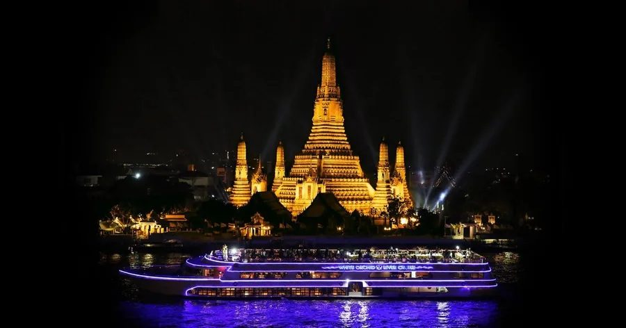Der Wat Arun nachts golden beleuchtet auf der anderen Flussseite, davor ein beleuchtetes Ausflugsschiff