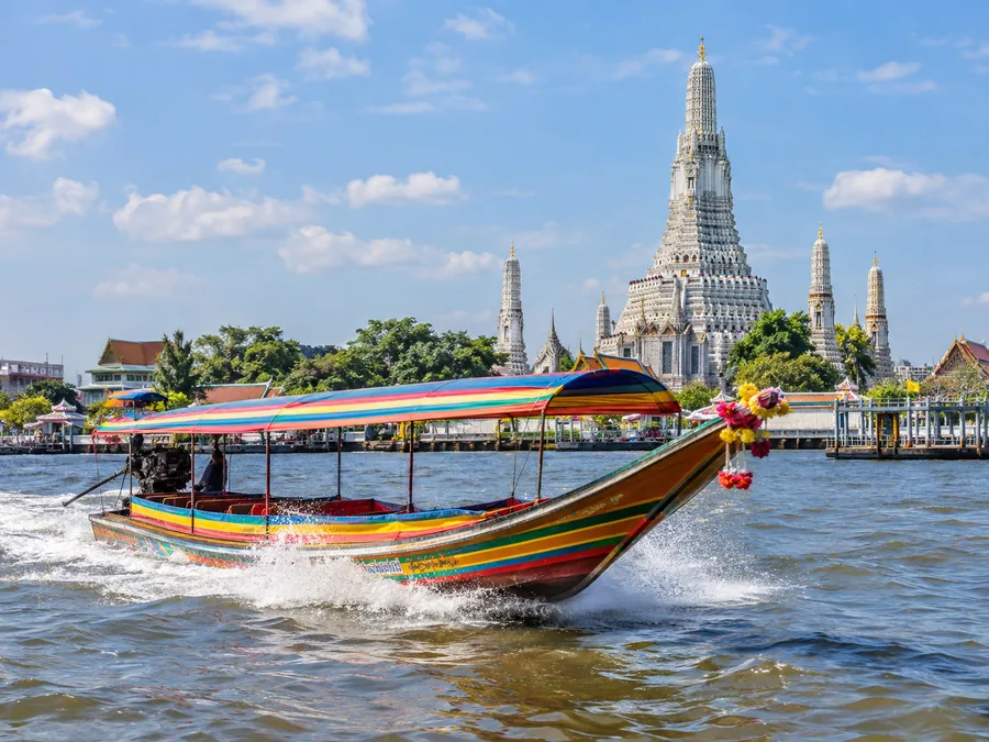 Ein hölzernes Longtail-Boot saust über den Chao Phraya, dahinter der weiße Prang des Wat Arun